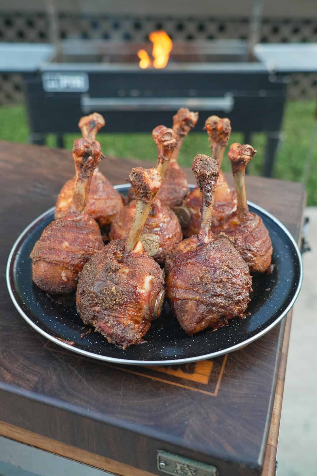 A plate of smoked honey BBQ bacon chicken lollipops in front of a grill fire