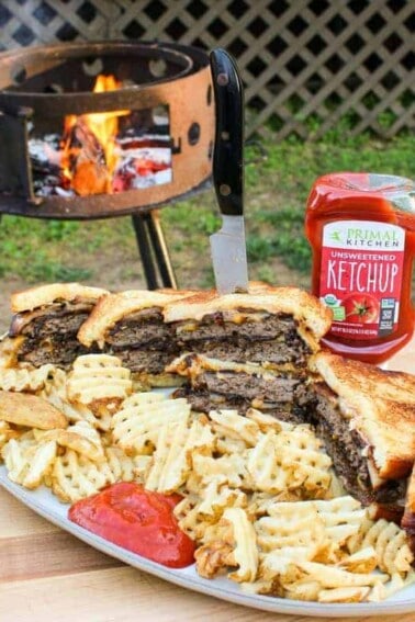 large platter on a table in front of a grill filled with cheesy patty melt sandwiches, fries, and ketchup