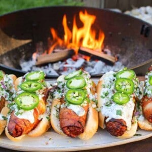 five bacon wrapped chorizo dogs topped with cream sauce and jalapeño in front of a fire pit