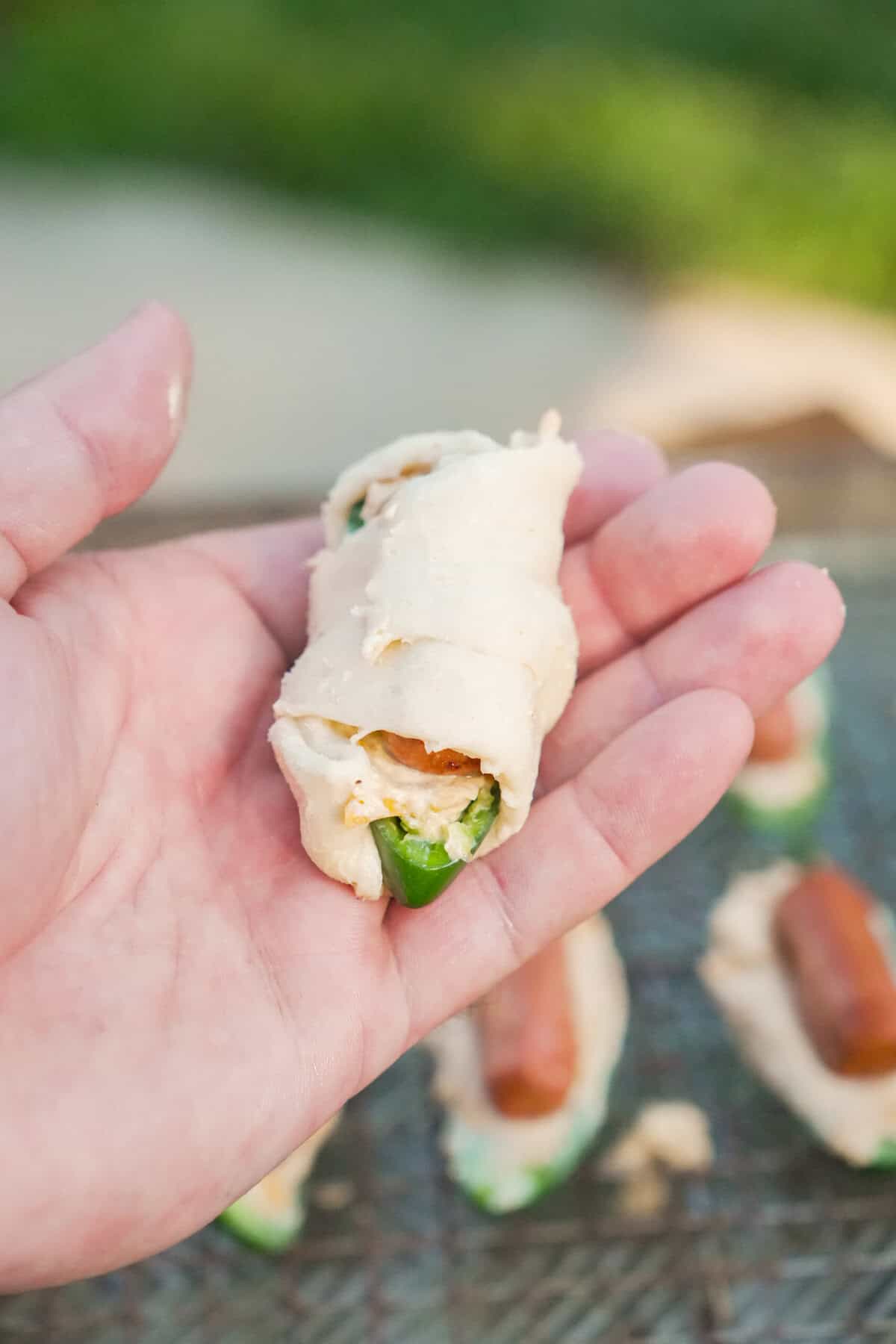 A hand holding a wrapped but uncooked jalapeño popper pig in a blanket