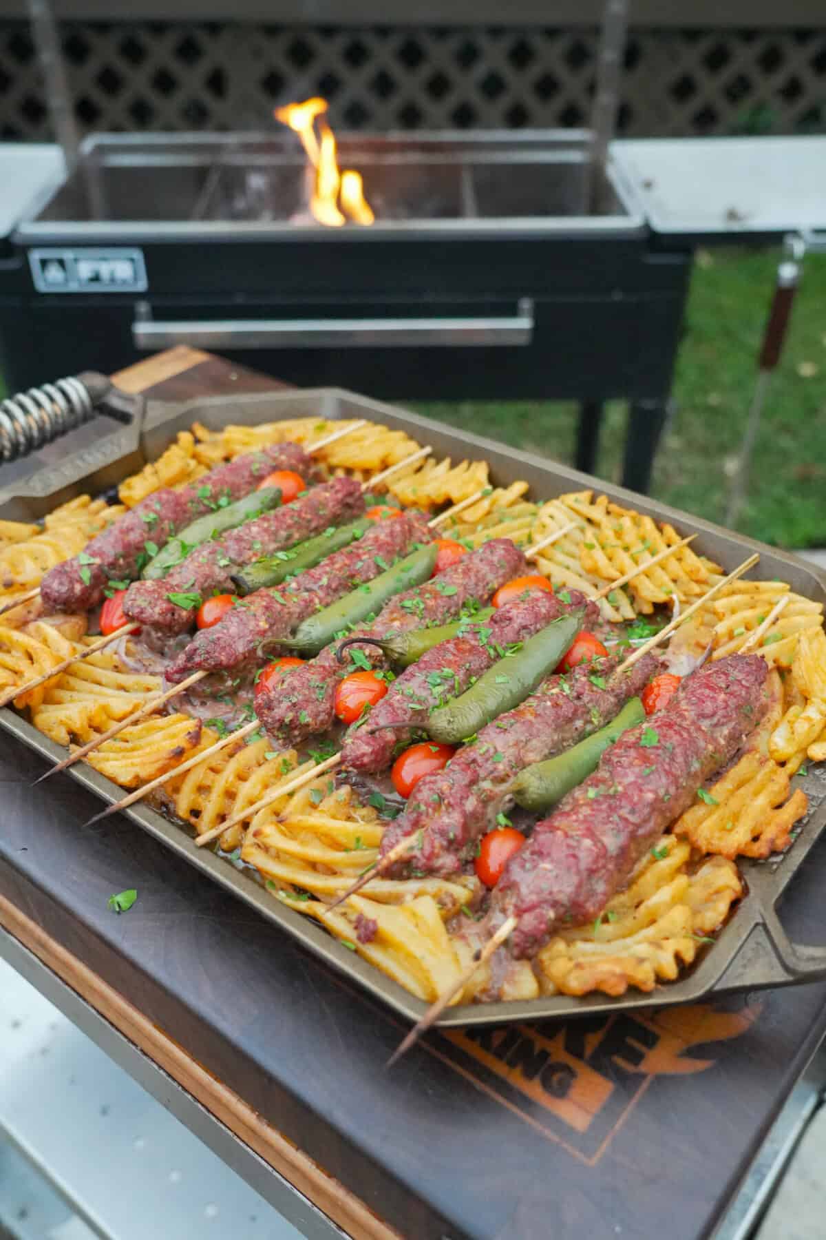 A platter of grilled beef kebabs with waffle fries, cherry tomatoes, sliced white onion and chile peppers