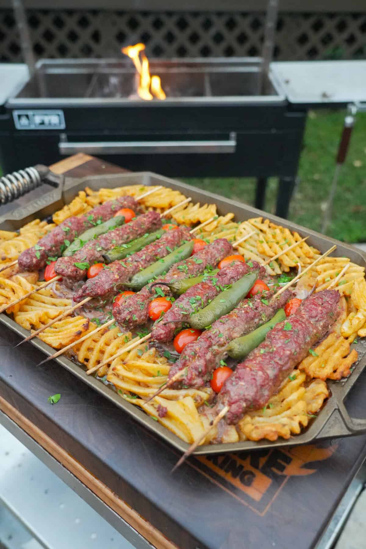 A platter of grilled beef kebabs with waffle fries, cherry tomatoes, sliced white onion and chile peppers