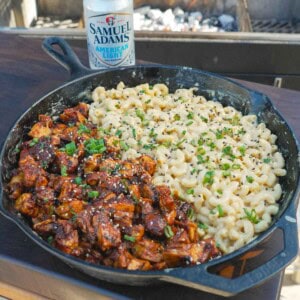 A cast iron skillet filled with BBQ chicken and beer cheese macaroni and a can of Samuel Adams in the background