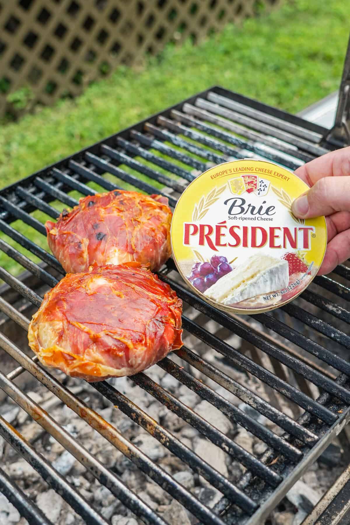 A hand holding a wheel of Président Brie cheese next to prosciutto-wrapped brie on the grill