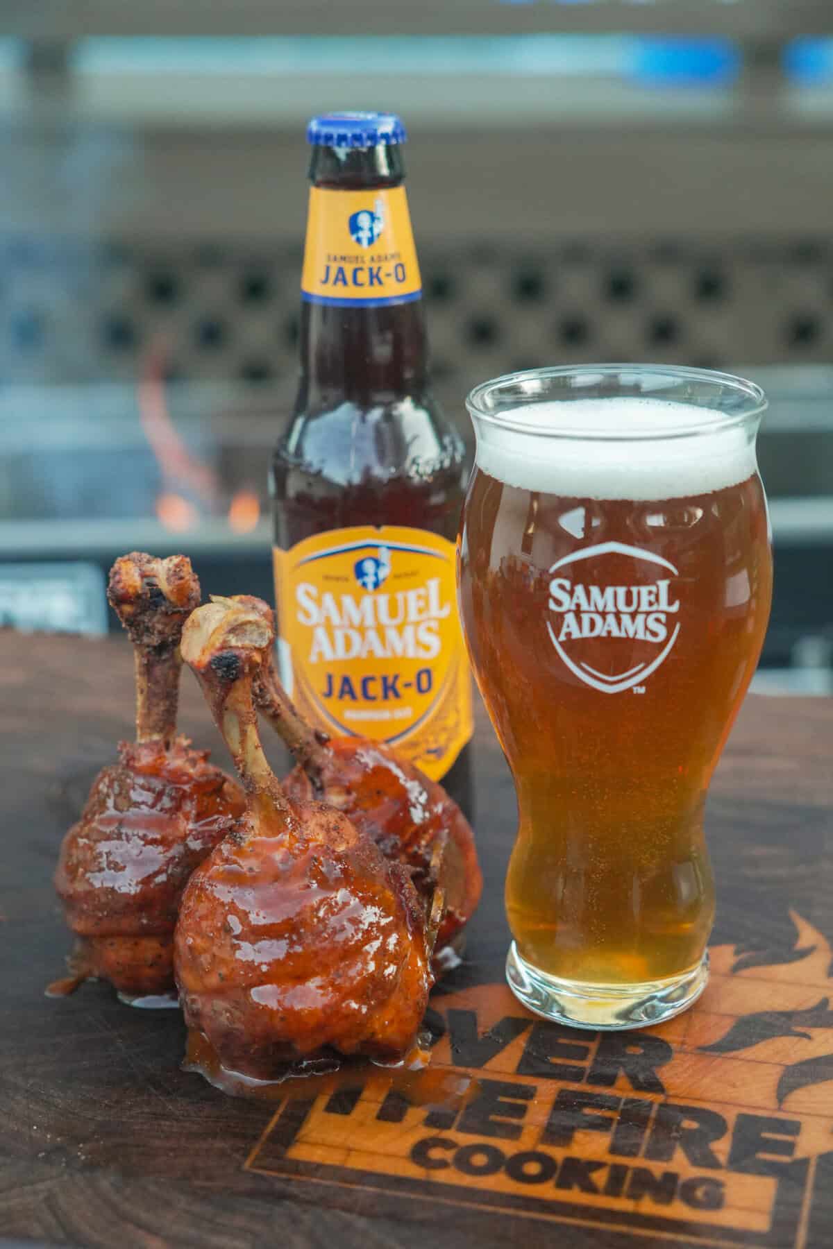 Two finished honey bacon BBQ chicken lollipops with a glass of Samuel Adams Jack-O in a glass and a bottle of the beer in the background