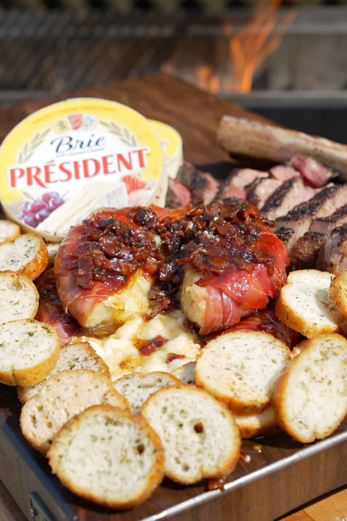 A close up of melty brie cheese wrapped in prosciutto with the Président container, crusty bread slices and slices of steak