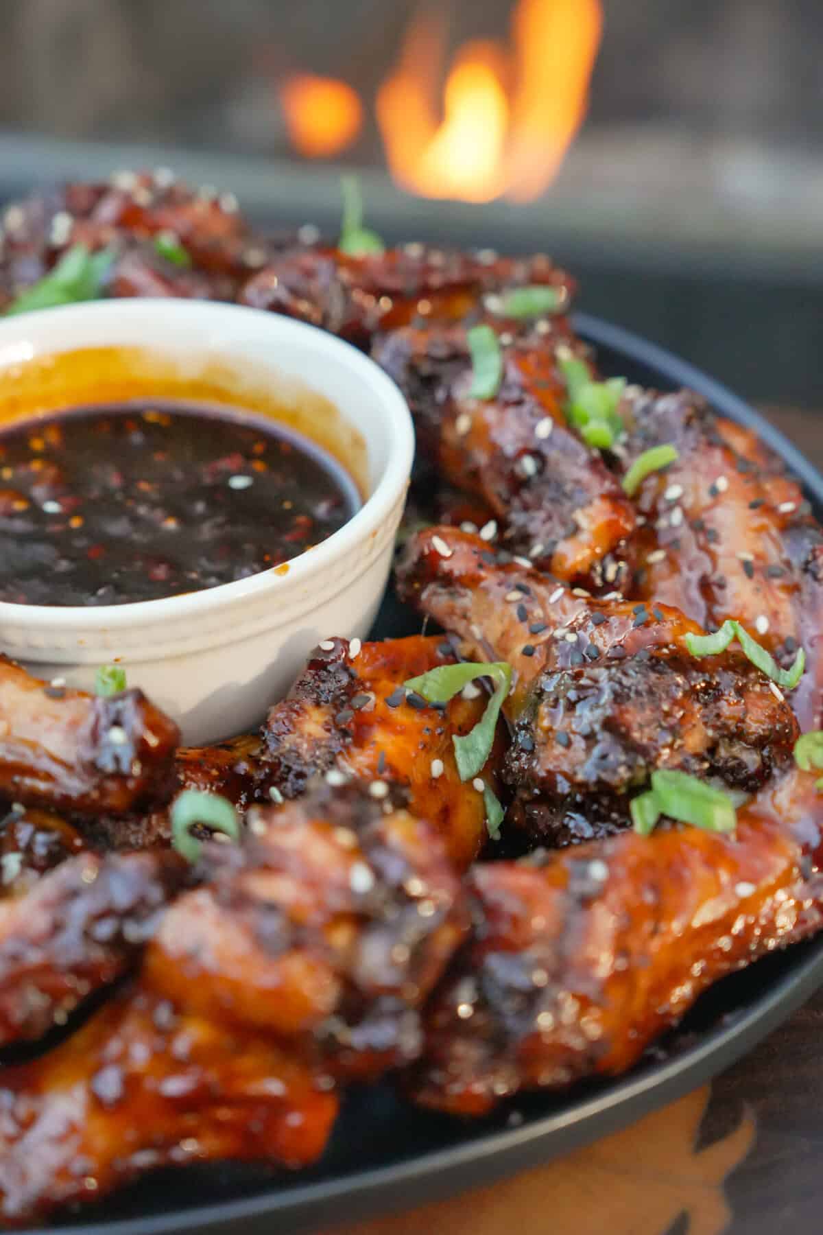 A close up of a platter of honey Korean gochujang wings with sesame seeds and scallions as garnish and dipping sauce in a bowl in the middle