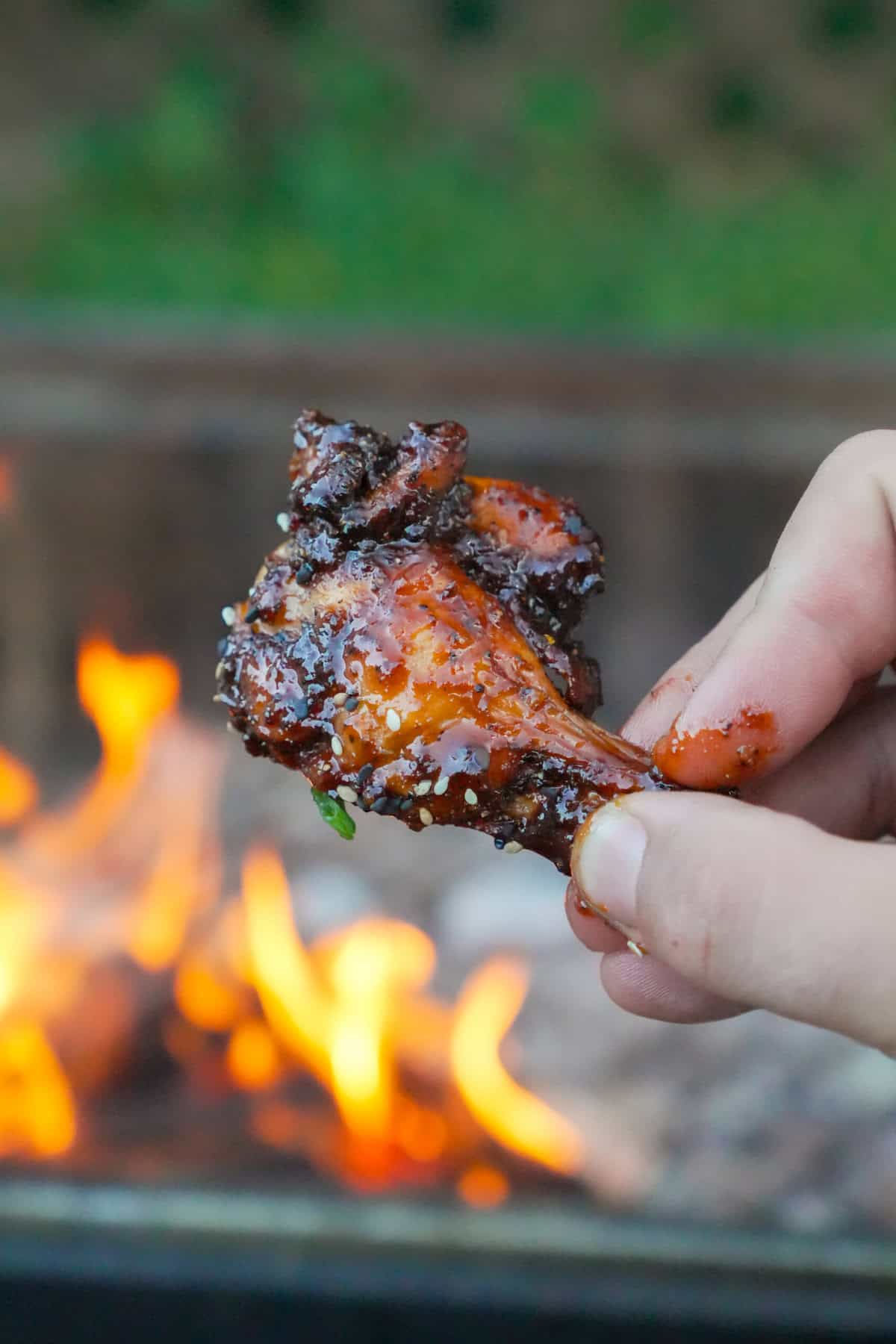A hand holding a grilled chicken wing with the grill fire in the background