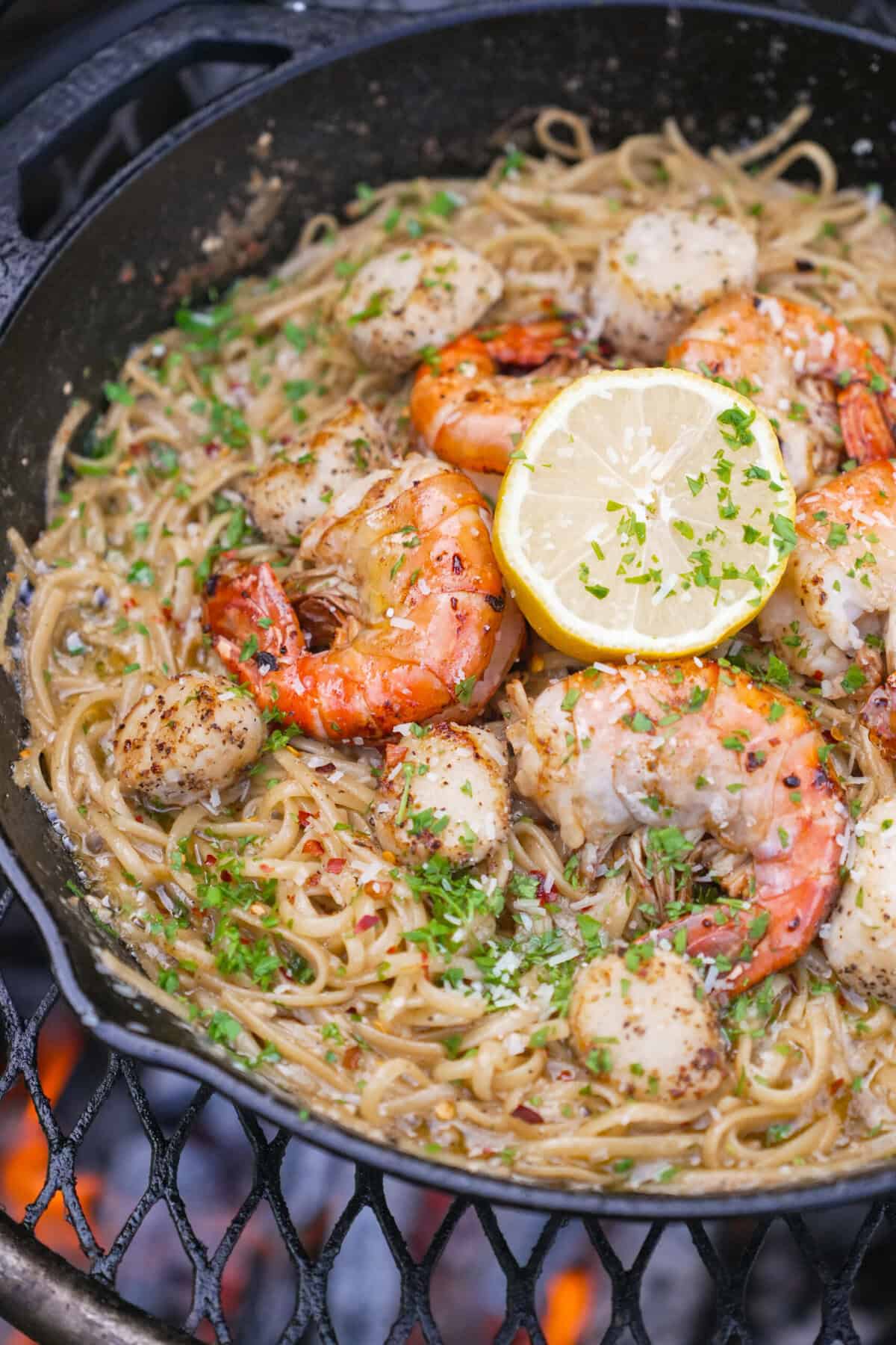 A close up shot of seafood scampi with pasta, shrimp, scallops and lemon in a cast iron skillet over an open fire