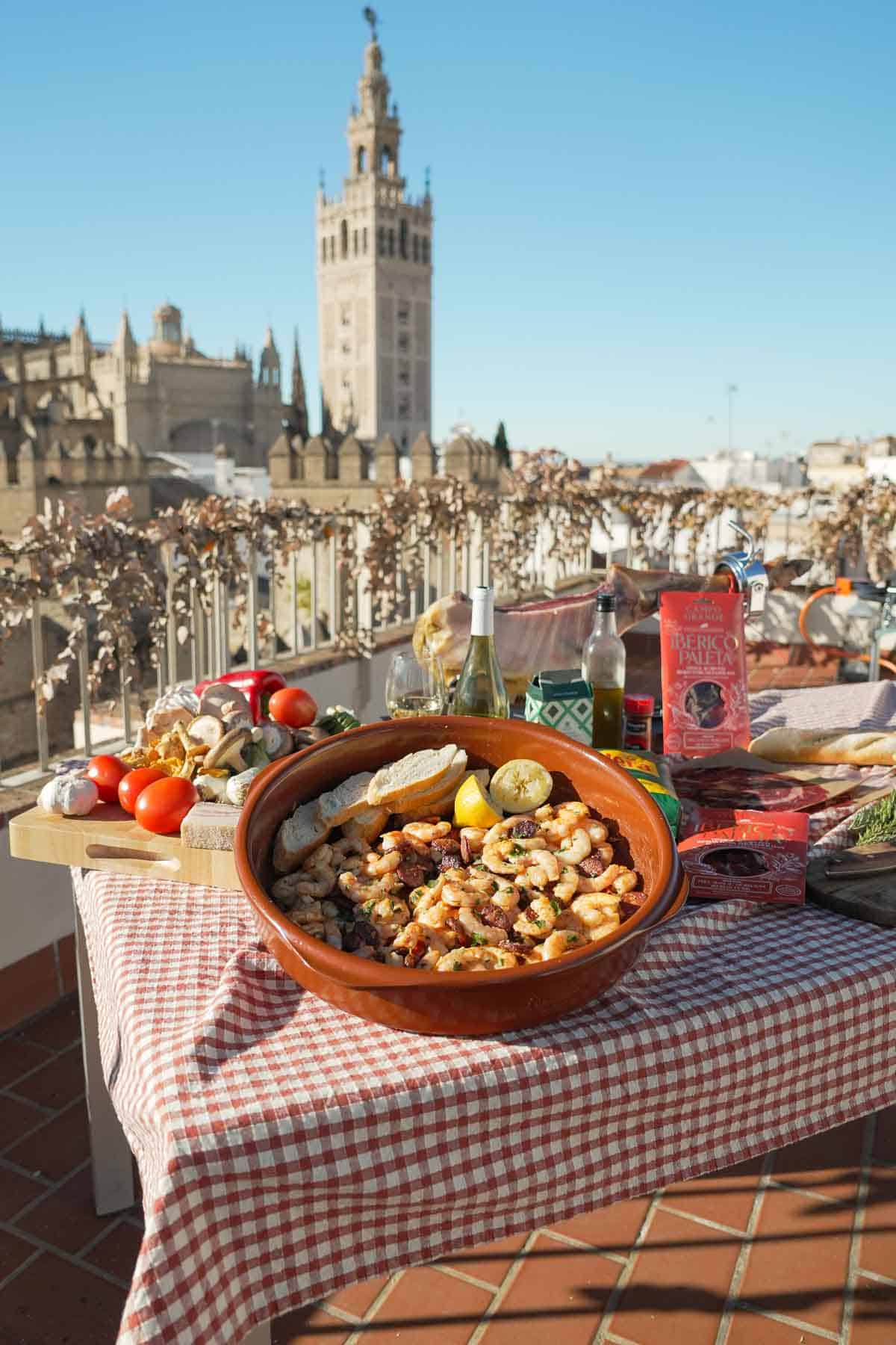 The dish is featured on a table shared with the fresh ingredients used in the recipe - with the beautiful historic city of Seville, Spain in the background.