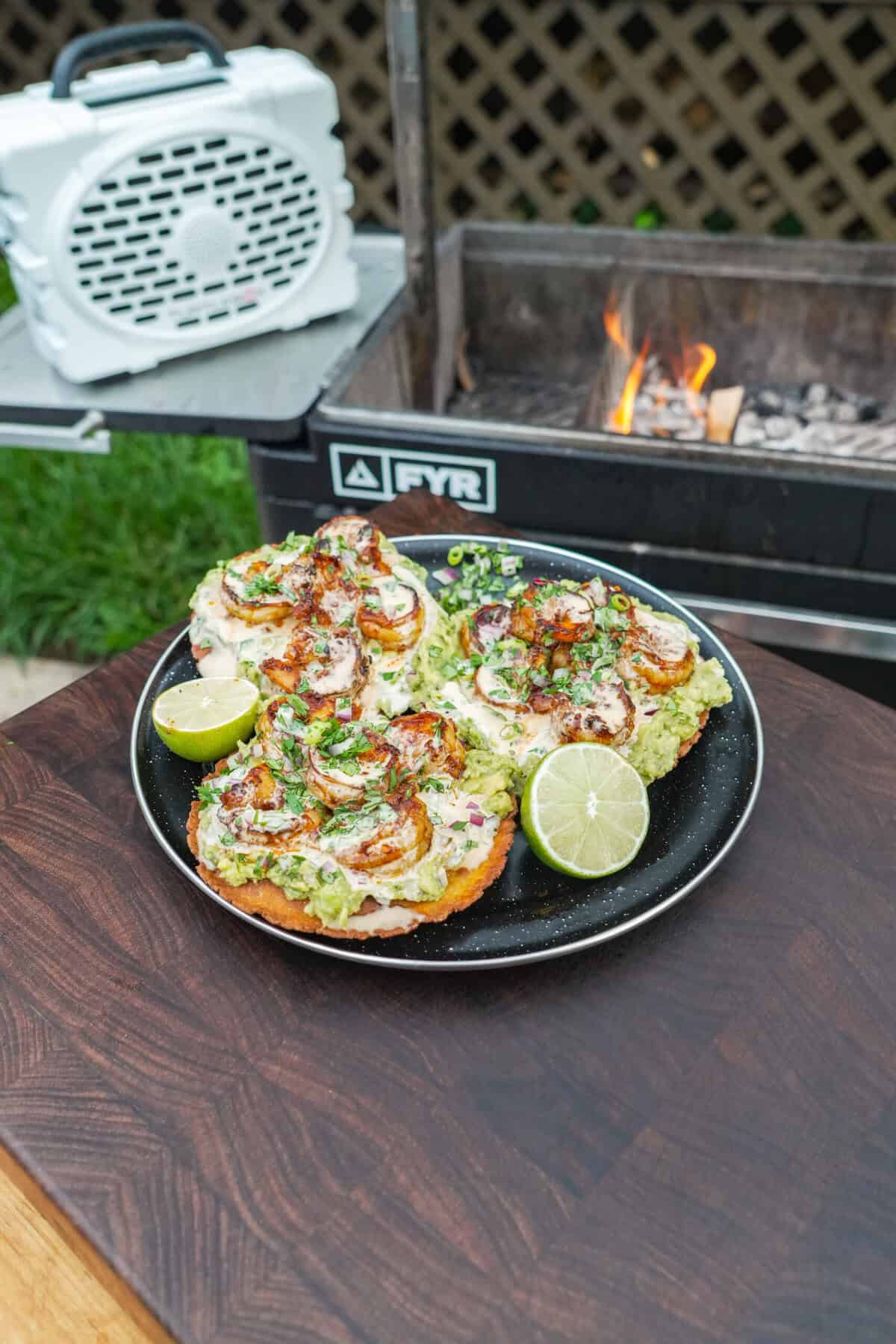 A plate of assembled shrimp tostadas with chili oil, guacamole and yum yum sauce, featuring a Turtlebox speaker and the FYR Grill in the background