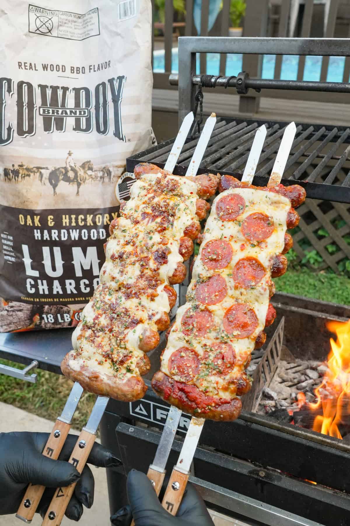 Two finished pizza sausage skewers — one pepperoni, one alfredo — standing next to each other in front of a FYR Grill and next to a bag of Cowboy Charcoal