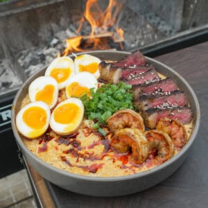 A full bowl of surf and turf ramen next to the grill, filled with steak, shrimp, eggs, noodles and garnishes