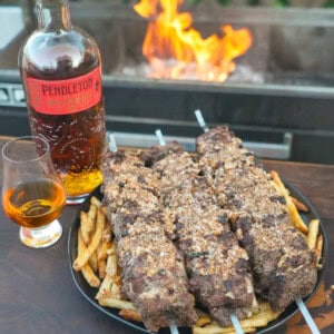 The French Onion Steak Skewers are plated next to a bottle of Pendleton Whisky next to the grill with flames.