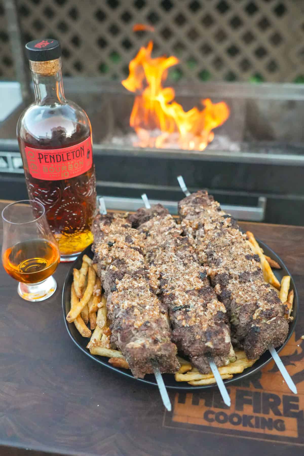 The French Onion Steak Skewers are plated next to a bottle of Pendleton Whisky next to the grill with flames. 