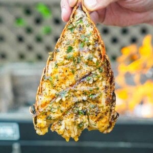 A hand holding a grilled lobster tail with honey Cajun butter and a grill in the background