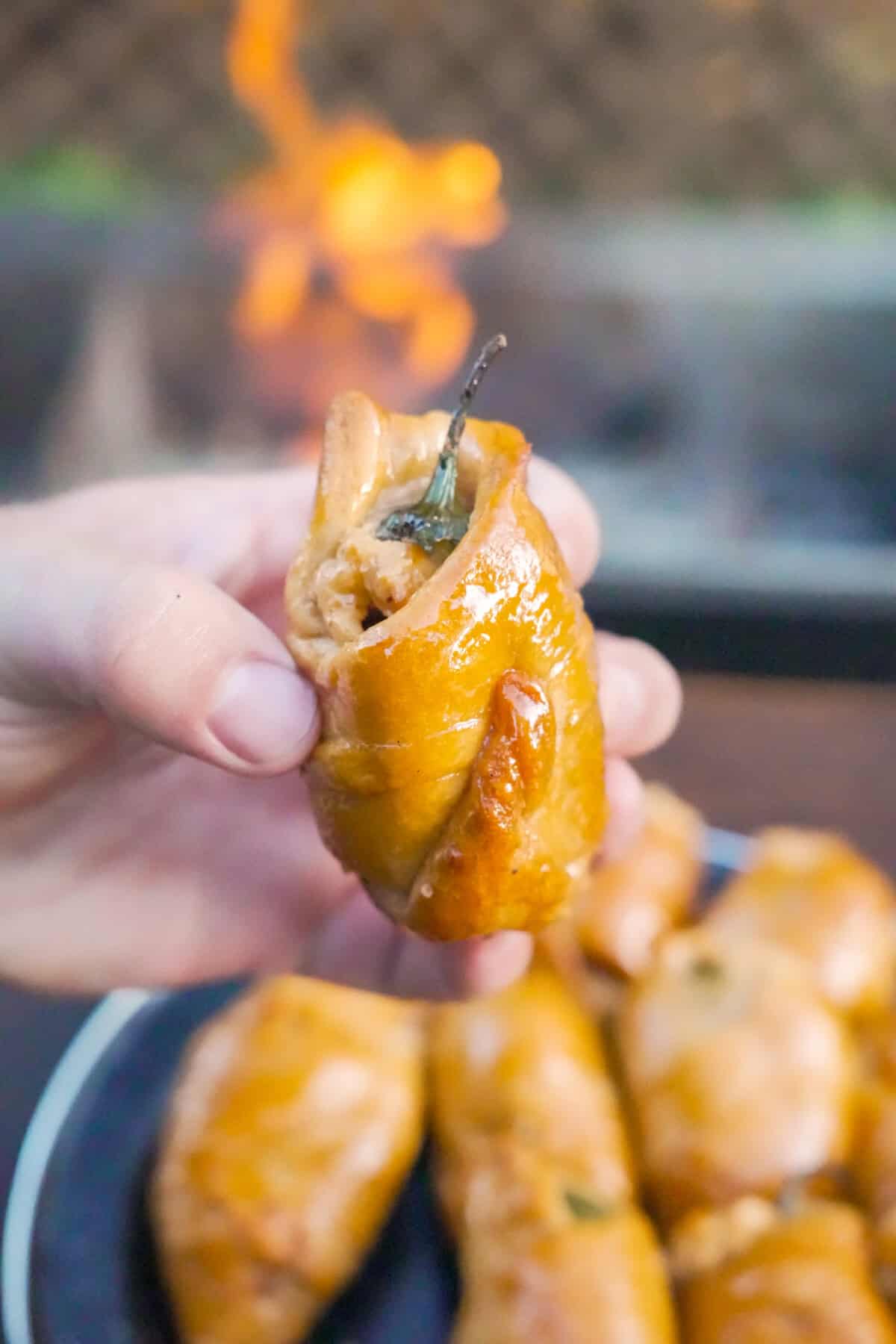 A hand holding one of the jalapeño popper pigs in a blanket, with a plate of the other pigs in the background and the grill fire