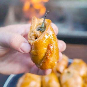 A hand holding one of the jalapeño popper pigs in a blanket, with a plate of the other pigs in the background and the grill fire