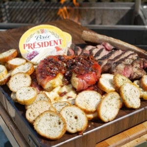 A plate filled with brie wrapped in prosciutto and topped with bacon jam, slices of crusty bread, slices of grilled steak and a container of Président Brie in the background