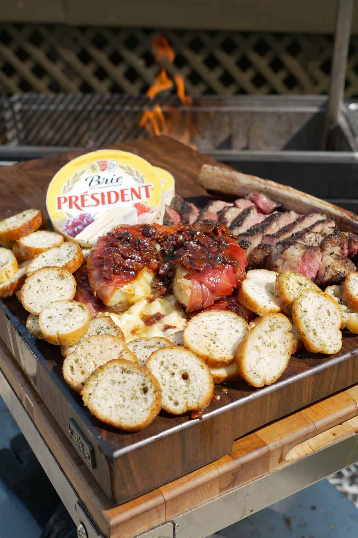 A plate filled with brie wrapped in prosciutto and topped with bacon jam, slices of crusty bread, slices of grilled steak and a container of Président Brie in the background