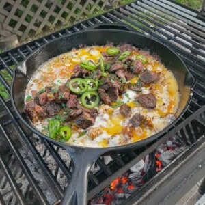 A cast iron skillet of queso flameado on the grill, with chopped cilantro and jalapeño slices for the garnish
