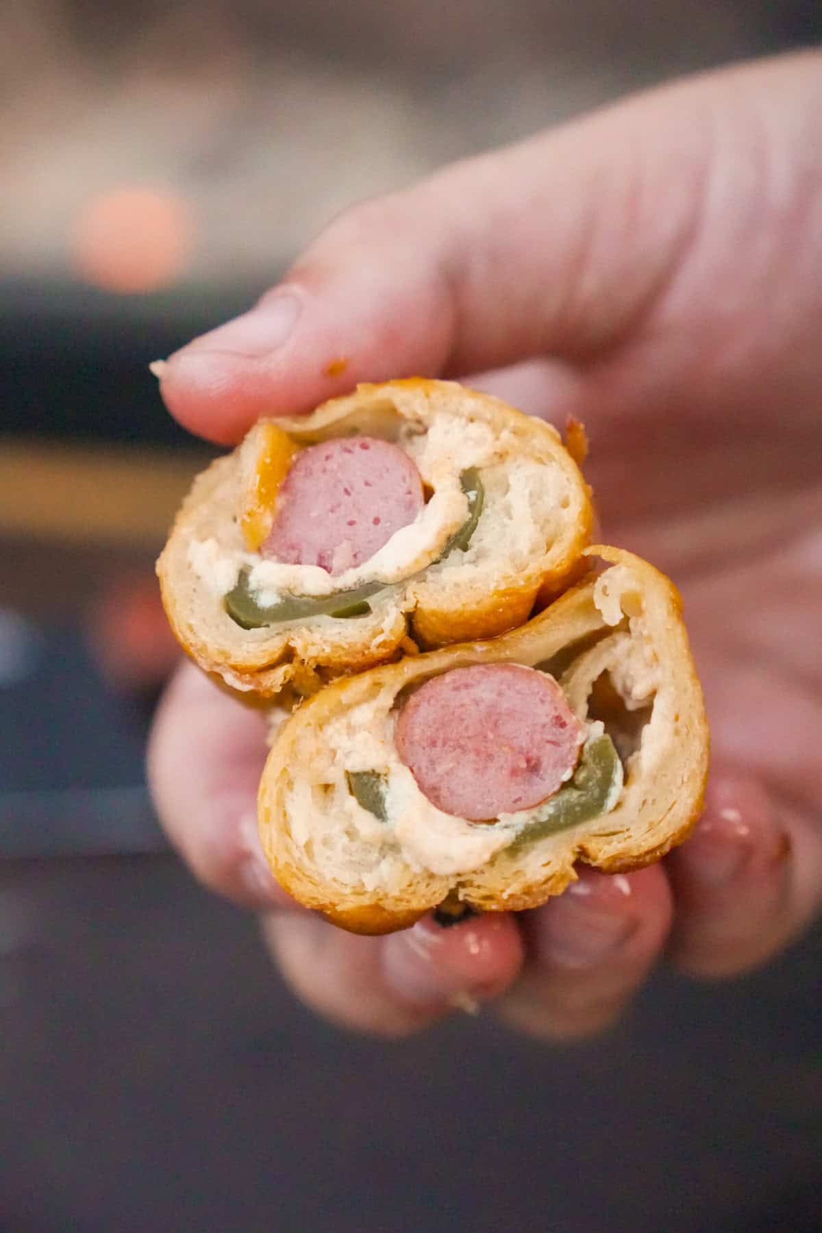 A hand holding a sliced open jalapeño popper pig in a blanket, showing the cocktail wiener, jalapeños and cheese spread