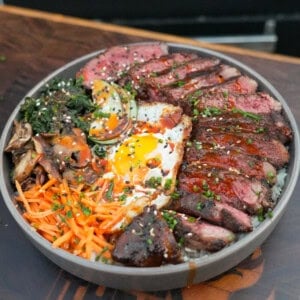 A finished bowl of steak bibimbap, with grilled ribeye, a runny fried egg, bibimbap sauce, carrots, mushrooms, spinach and cucumbers